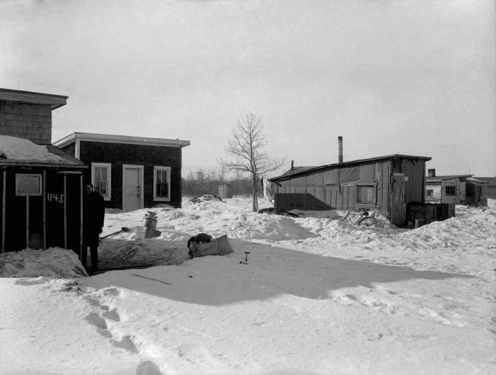 Gerry Cairns / Winnipeg Free Press Collection, Archives of Manitoba
A news reporter stands outside the home of James and Mary Parisien at 1145 Weatherdon Avenue in 1959. Despite their simple construction, the shanties displayed some variations in status. The wooden shingle siding of the Parisien residence distinguishes it from the tar-paper cladding of some of its neighbours. Beside 1145 Weatherdon is a home with painted trim and sashes.