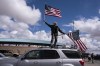 Jeff Cassil waves two American flags from the roof of his car at the beginning of a trucker caravan to Washington, D.C., called The People's Convoy Wednesday, Feb. 23, 2022, in Adelanto, Calif. A small convoy of truckers demanding an end to coronavirus mandates began a cross-country drive from California to the Washington, D.C., area on Wednesday. (AP Photo/Nathan Howard)